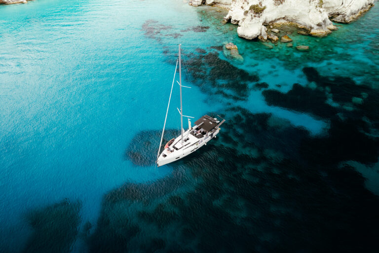 Ein Segelboot ankert in einer ruhigen, türkisfarbenen Bucht, umgeben von teilweise unter Wasser liegenden Felsen und weißen Klippen – perfekt für eine ruhige Pause während einer Atlantiküberquerung. Das Wasser variiert in Blautönen, mit Flecken von dunklerem Blau und Grün um das Boot herum.