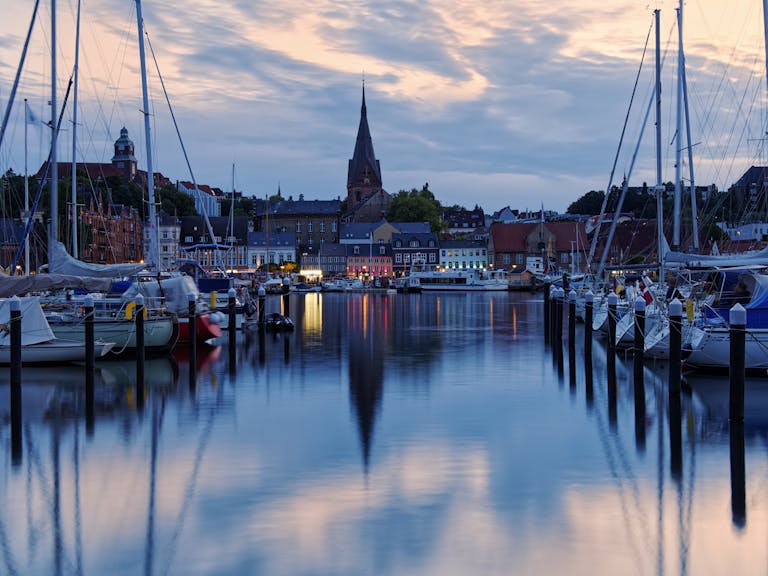 Peaceful evening view of Flensburg Harbor, Germany, with moored sailboats and illuminated waterfront buildings.